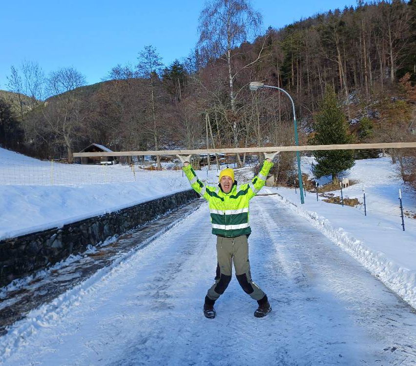 Ein Mann in einer gelben und grünen Jacke hält einen langen Holzbalken, während er im Schnee mit gespreizten Beinen steht. Dahinter befindet sich eine Steinmauer, eine Straßenlaterne und ein kleines Haus.