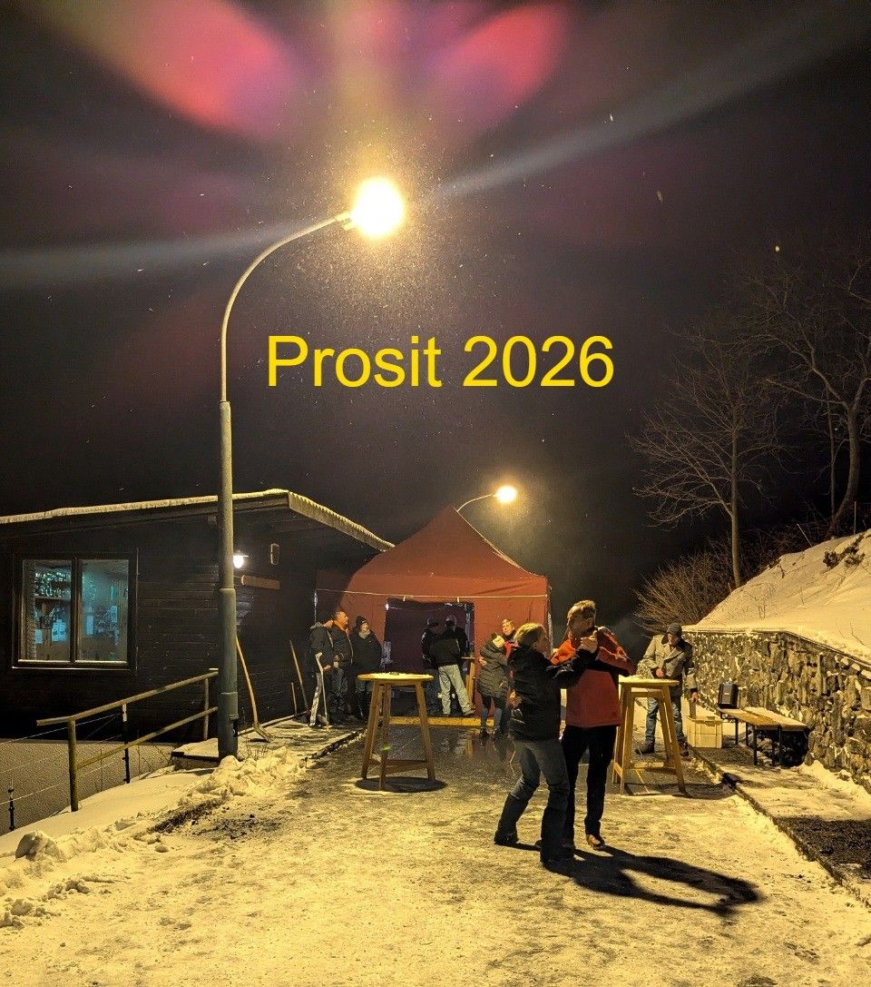 In der Nacht tanzen Menschen vor einem Zelt mit der Aufschrift Prosit 2026. Eine Straßenlaterne steht links, und ein Gebäude befindet sich rechts.