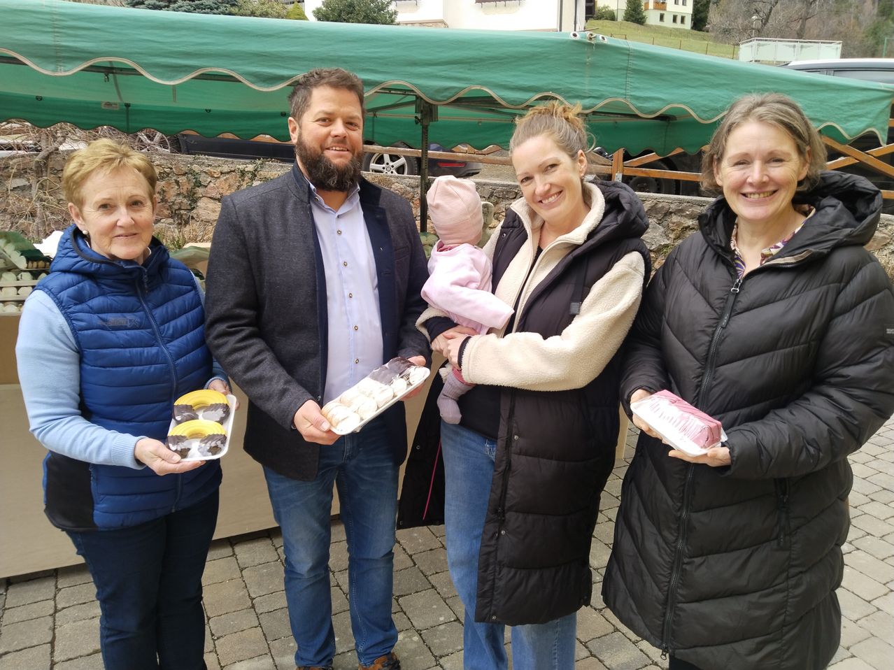 Vier Erwachsene, zwei mit Desserts und eine Frau mit einem Baby, lächeln unter einem grünen Zelt. Sie sind draußen in der Nähe einer Steinmauer.