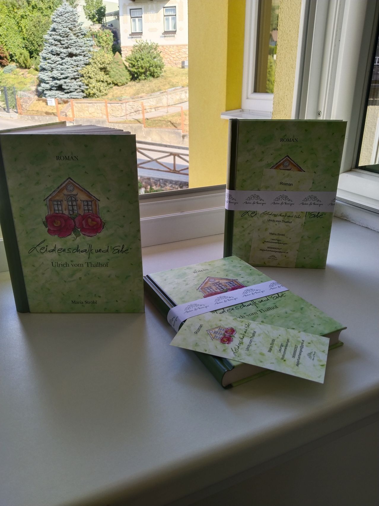 Drei Bücher mit den Titeln 'Keiderschaf und Elfe' und 'Roman' sind vor einem Fenster auf einer weißen Oberfläche angeordnet. Die Bücher haben Illustrationen von Häusern und Blumen auf ihren Umschlägen.