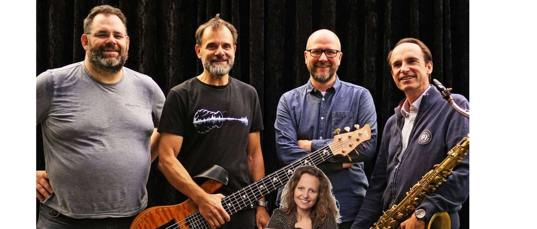 Fünf Musiker stehen zusammen für ein Foto. Vier Männer und eine Frau. Sie alle lächeln. Drei der Männer halten Instrumente. Einer hat eine Saxophon, der andere eine Gitarre. Die Frau sitzt auf einem Stuhl in der Mitte.
