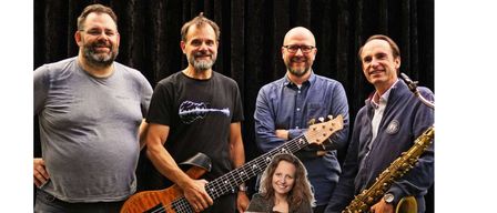 Five musicians are standing together for a photograph. Four men and one woman. They are all smiling. Three of the men are holding instruments. One has a saxophone, another a guitar. The woman is sitting on a chair in the center.
