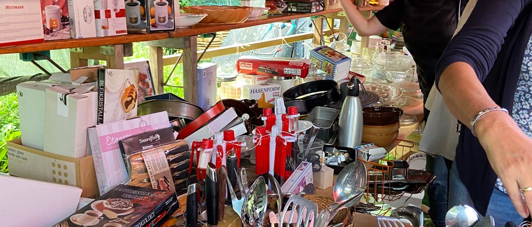Zwei Frauen durchstöbern einen Marktstand mit verschiedenen Artikeln wie Besteck, Schüsseln und Kartons. Eine Frau hält eine weiße Schüssel.