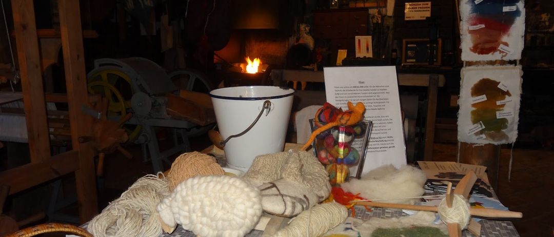 Ein Workshop mit verschiedenen Wollprodukten. Ein weißer Eimer, Garnknäuel und handgefertigte Wollartikel auf einem Tisch. Ein Kamin mit einem brennenden Feuer im Hintergrund.
