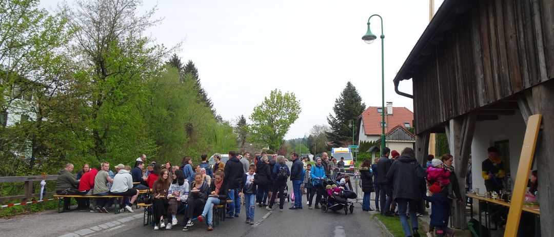 Eine Menschenmenge versammelt sich im Freien, einige sitzen auf Stühlen, andere stehen. Ein Krankenwagen ist in der Nähe geparkt. Bäume säumen die Straßenseite.