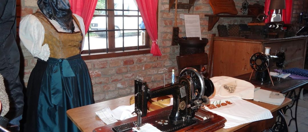Eine alte Nähmaschine auf einem Holztisch in einem Raum mit Backsteinwänden, roten Vorhängen und verschiedenen Gegenständen. Eine Person in traditioneller Kleidung steht in der Nähe.