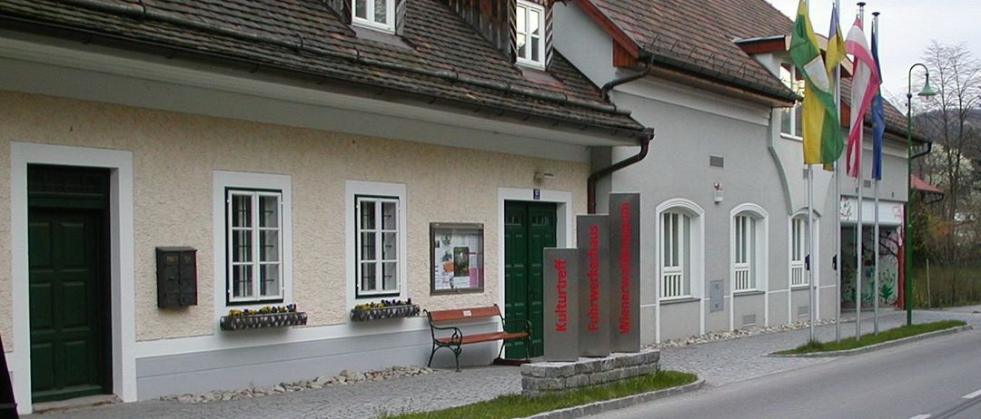Ein traditionelles Gebäude mit einem braunen Dach und weißen Fenstern. Ein Bank und ein Schild stehen davor. Auf der rechten Seite flattern Flaggen.