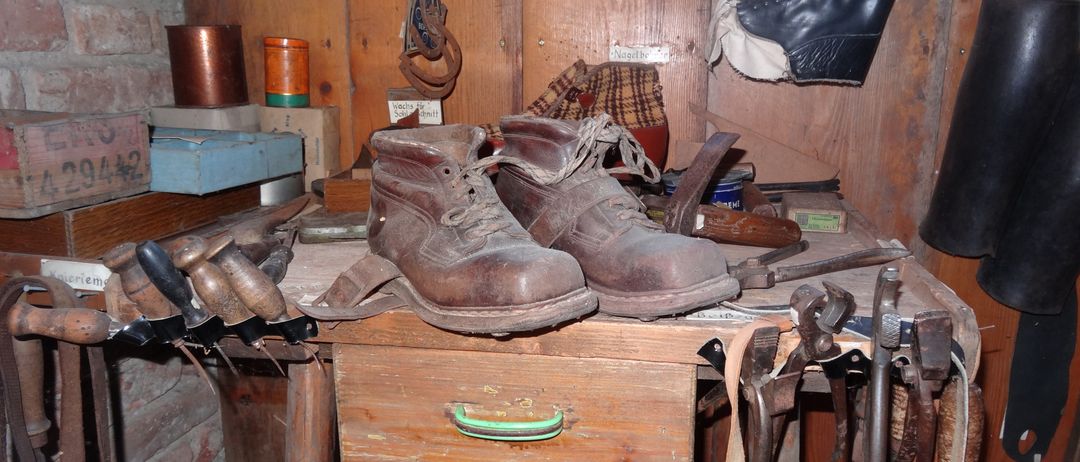 Ein Paar alte Lederstiefel steht auf einem Holzarbeitstisch, umgeben von verschiedenen Werkzeugen und Gegenständen, darunter eine Dose, ein Kasten und ein Hufeisen.