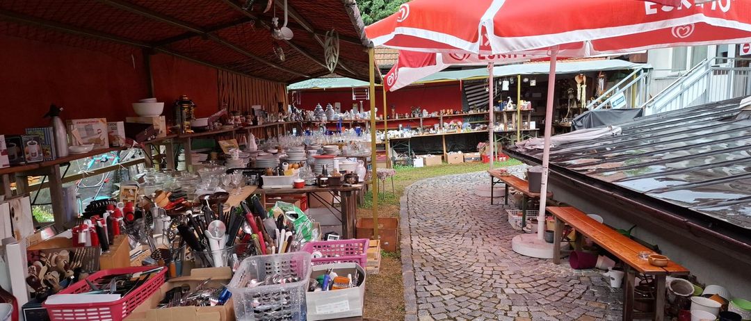 Ein Flohmarkt mit verschiedenen Artikeln unter roten Sonnenschirmen, einschließlich Küchenutensilien, Tellern und Schüsseln, entlang eines Kopfsteinpfades.