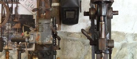 Eine Werkstatt mit verschiedenen alten Maschinen und Werkzeugen, einschließlich einer Bohrmaschine und einer Schweißmaske, an einer Steinwand montiert.