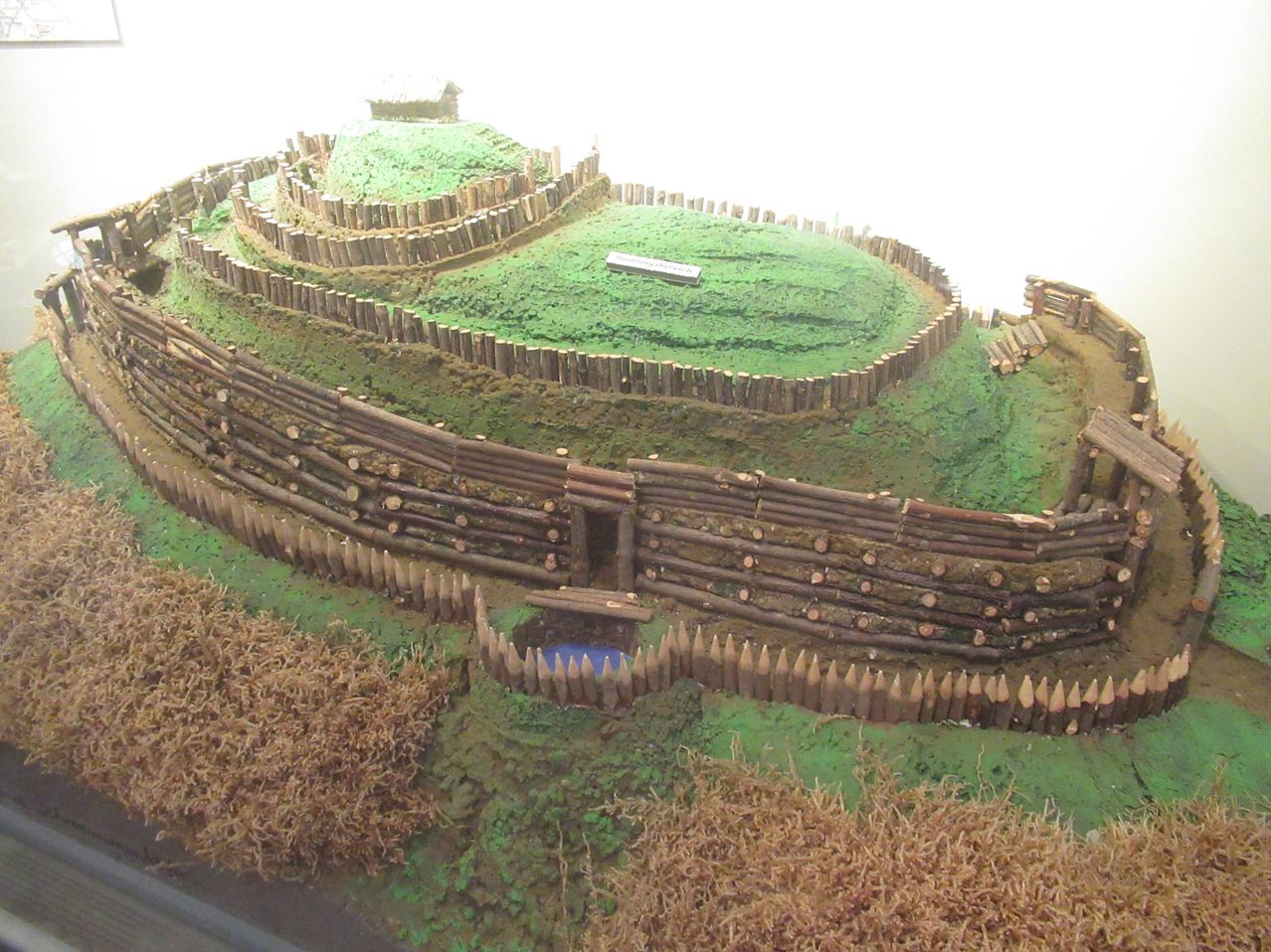 Ein Miniaturmodell einer Holzfestung mit einer spiralförmigen Befestigungsanlage und einem kleinen Holzhaus oben. Die Festung ist von einem grünen Feld und einem kleinen Teich umgeben.