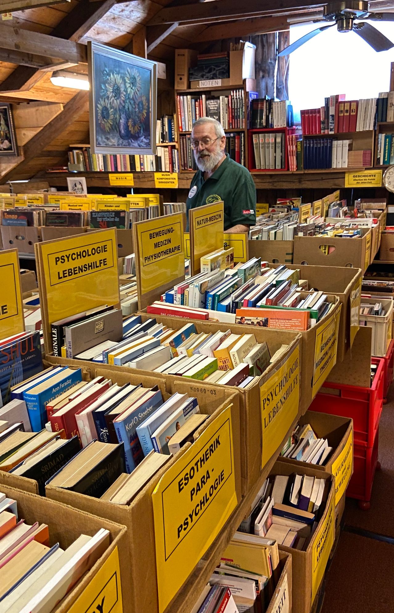Ein Mann in einem grünen Shirt steht in einem Raum voller Bücher, einige in Kisten mit den Aufschriften 'Psychologie' und 'Lebenshilfe'. Im Hintergrund befinden sich weitere Bücherregale.