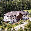 Ein Luftbild eines großen, weißen Gebäudes mit einem grauen Dach, eingebettet zwischen grünen Bäumen und einem Grasfeld. Das Gebäude hat mehrere Fenster und eine Terrasse mit grünen Sonnenschirmen.