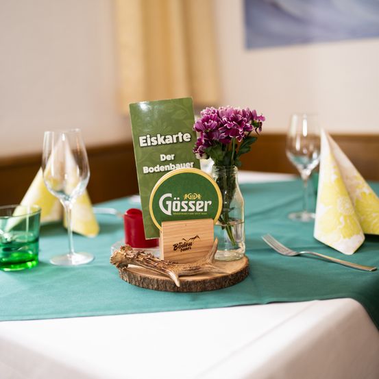 Eine Tischdekoration mit grüner Tischdecke, Weingläsern, einer Blumenvase und einem Holzschild mit dem Wort 'Gösser'.