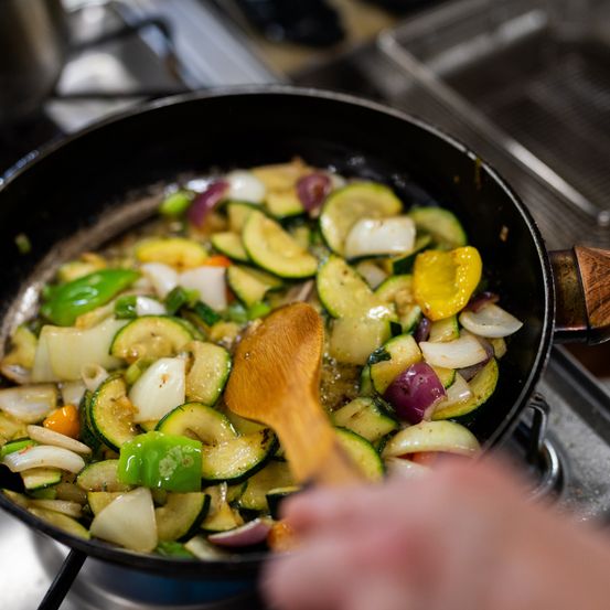 Ein hölzerner Löffel rührt eine Pfanne mit gemischtem Gemüse, darunter Zucchini, Paprika und Zwiebeln, auf einem Herd.