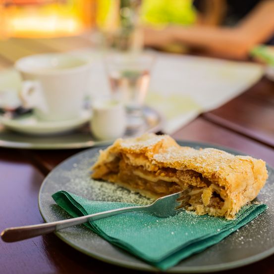 Ein Stück Apfelstrudel auf einem Teller mit einer Gabel, bestreut mit Puderzucker, begleitet von einer Tasse Kaffee.