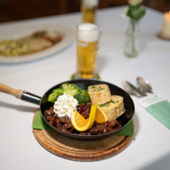 Ein Nahaufnahme eines Pfannengerichts mit rotem Fleisch, Orangenscheiben und grünem Gemüse auf einem Holzteller, begleitet von einem Glas Bier und einer Kerze.