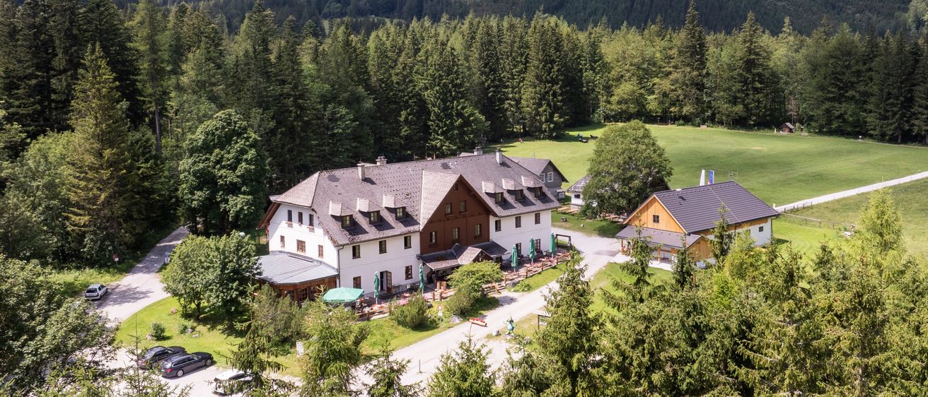 Ein Luftbild eines großen Chalets, umgeben von einem üppigen grünen Feld und dichtem Wald. Das Chalet hat ein graues Dach und braune Wände. Vor dem Chalet stehen mehrere grüne Sonnenschirme.