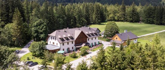 Ein Luftbild eines großen Chalets, umgeben von einem üppigen grünen Feld und dichtem Wald. Das Chalet hat ein graues Dach und braune Wände. Vor dem Chalet stehen mehrere grüne Sonnenschirme.