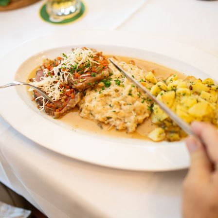 Ein Teller mit traditionellem deutschen Essen mit Fleisch, Kartoffeln und Gemüse, garniert mit Käse und Kräutern, wird mit einem Messer geschnitten.