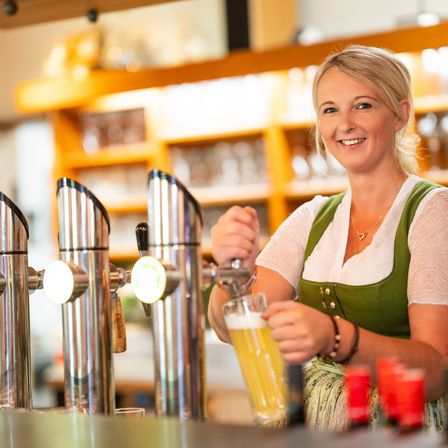 Eine Frau in einem grünen Kleid lächelt, während sie ein Glas Bier aus einem Zapfhahn in einer Bar schenkt.