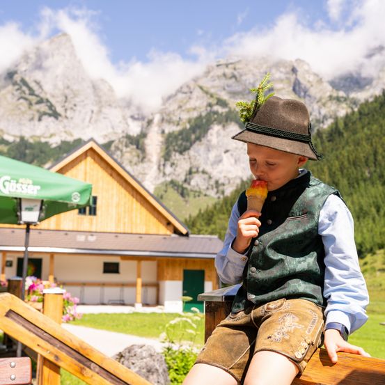 Ein junger Junge in traditioneller bayerischer Kleidung isst einen Eisbecher vor einer Bergkulisse.