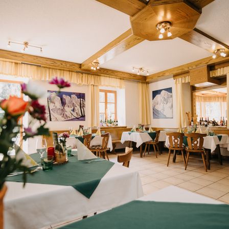 Ein leeres Restaurant mit Holzstühlen und Tischen, geschmückt mit weißen und grünen Tischdecken. Die Wände sind mit Berggemälden verziert, und die Decke hat Holzbalken und Beleuchtungskörper.