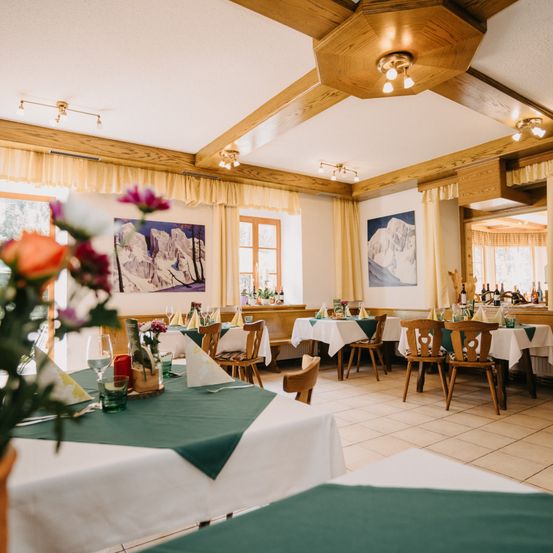 Ein leeres Restaurant mit Holzstühlen und Tischen, geschmückt mit weißen und grünen Tischdecken. Die Wände sind mit Berggemälden verziert, und die Decke hat Holzbalken und Beleuchtungskörper.