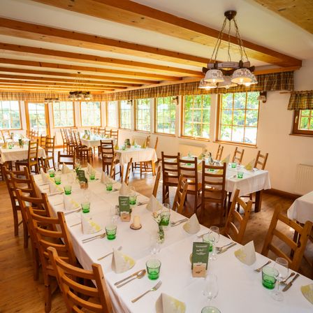 Ein Speisesaal mit mehreren langen Tischen, die für eine Mahlzeit gedeckt sind, mit Holzstühlen, weißen Tischdecken und grünem Glas. Holzbalken und große Fenster mit Vorhängen schmücken den Raum.