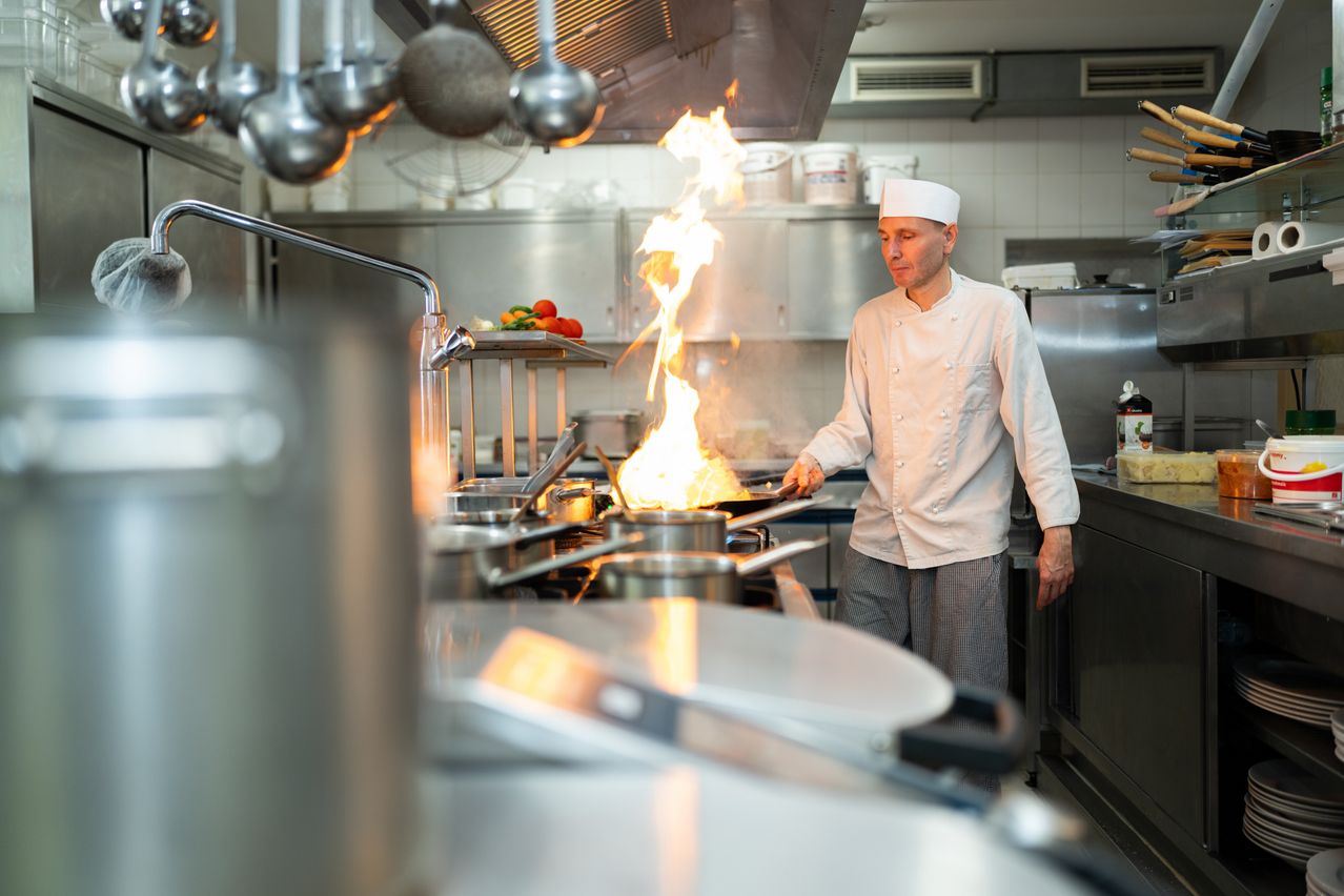 Ein Koch kocht in einer Küche, aus der Pfanne, die er hält, kommt Feuer.
