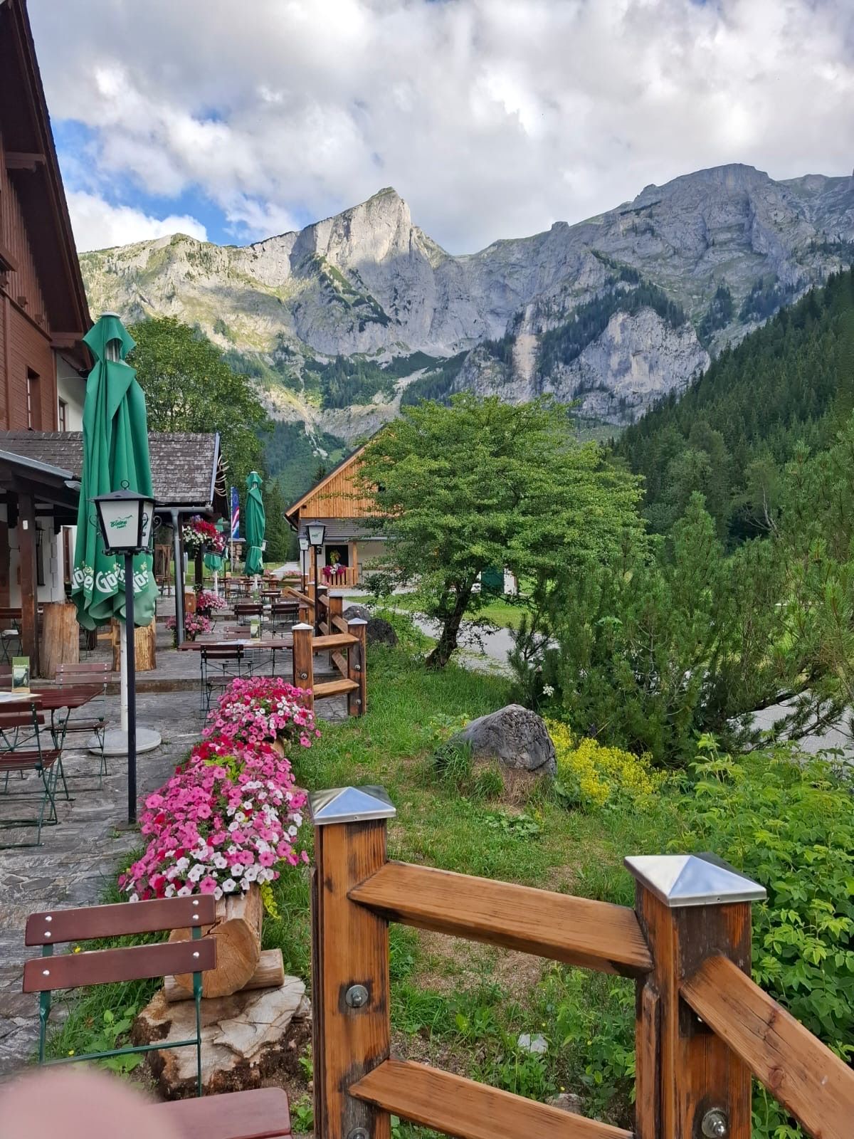 Ein malerisches Panorama eines Bergdorfes mit einer Restaurantterrasse, farbenfrohen Blumen und einem Holzzaun vor der Kulisse majestätischer Berge.