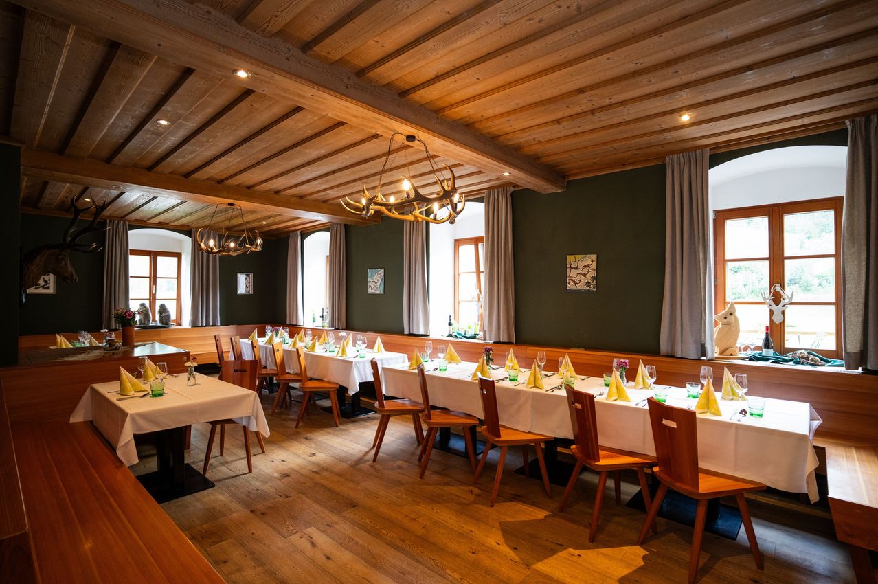 An empty dining hall with wooden floors, wooden chairs, and long tables set with white tablecloths and yellow napkins. The room has wooden beams, green walls, and chandeliers.