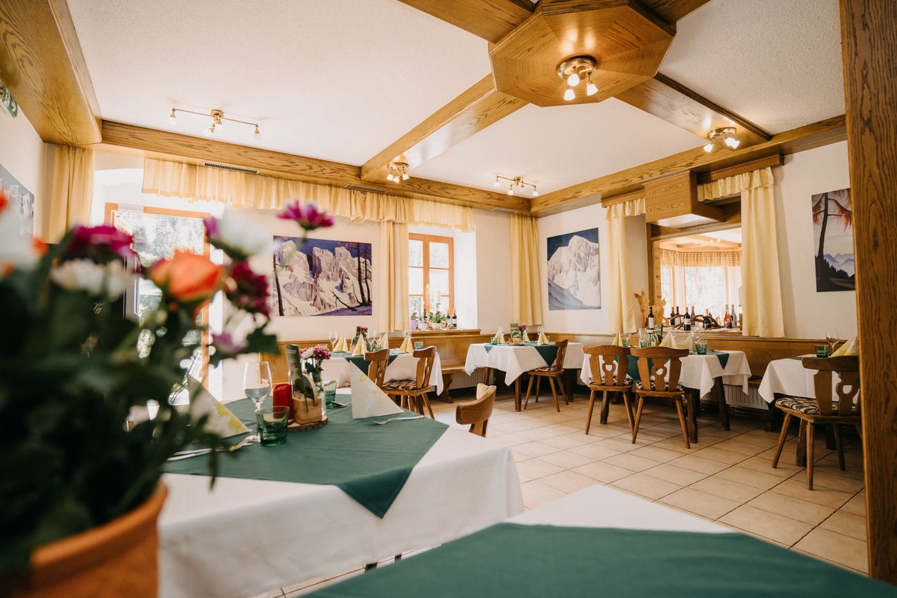 A well-decorated restaurant with wooden furniture, floral centerpieces, and scenic paintings on the walls. The room is bright with natural light and has a cozy atmosphere.