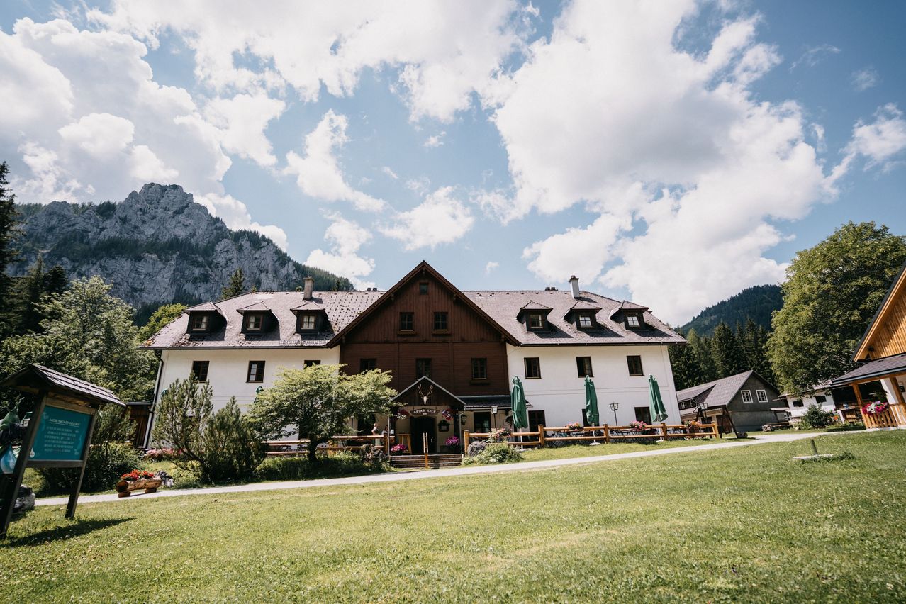 Ein großes, braunes und weißes Chalet-Stil-Gebäude mit mehreren Fenstern und einem grauen Dach steht vor einem grasbewachsenen Feld unter einem teilweise bewölkten Himmel, mit Bergen im Hintergrund.