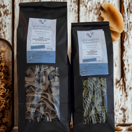 Zwei schwarze Tüten mit Nudeln sind ausgestellt. Eine Tüte enthält Bandnudeln Stemplitz, während die andere Radiatore Baratchi enthält. Beide sind in einer hölzernen Umgebung aufbewahrt.