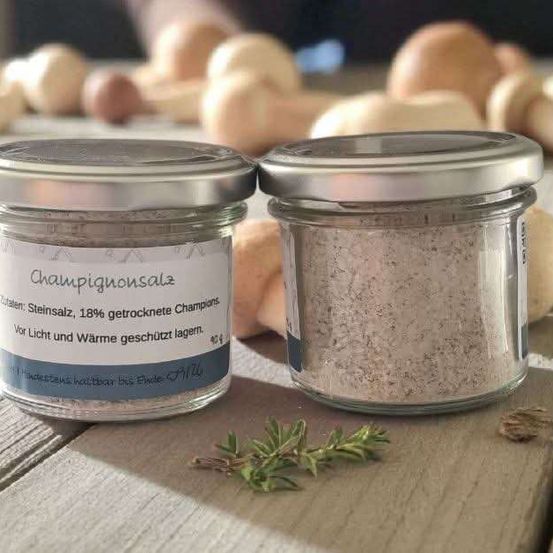 Zwei Glasgläser mit Deckeln auf einem Holztisch, eines mit der Aufschrift 'Champignonsalz', das getrocknete Pilze enthält, und ein weiteres mit weißem Pulver. Dahinter sind mehrere ganze Pilze zu sehen.