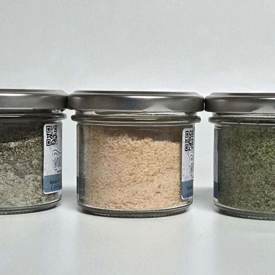 Drei Glasgläser mit silbernen Deckeln, gefüllt mit verschiedenfarbigen Pulvern, mit QR-Codes beschriftet. Die Gläser stehen nebeneinander auf einer weißen Oberfläche.