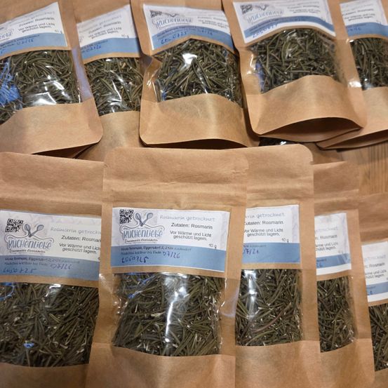 Mehrere braune Papiertüten enthalten getrocknete Rosmarinkräuter, mit blauen Etiketten, die den Markennamen 'Kuchenliebe' anzeigen. Der Rosmarin ist versiegelt, um die Frische zu bewahren, und die Etiketten enthalten auch einen QR-Code.