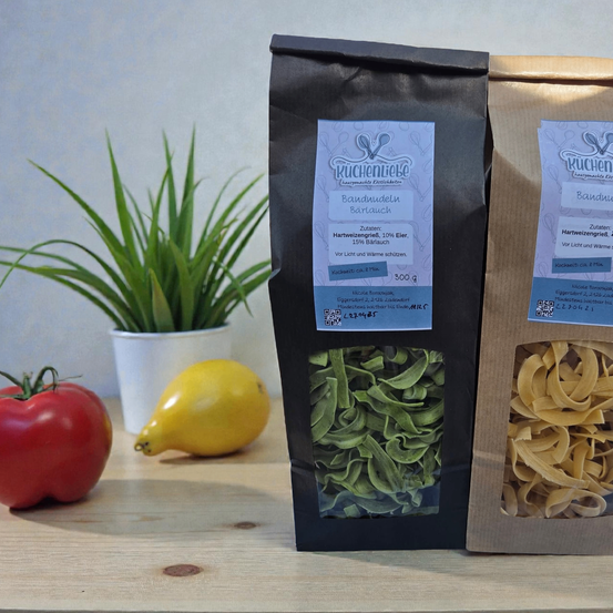 Zwei Tüten Pasta stehen auf einem Holztisch mit einer Tomate, einer Birne und einem Topf. Die Pasta hat Etiketten mit Zutaten und Nährwertangaben.