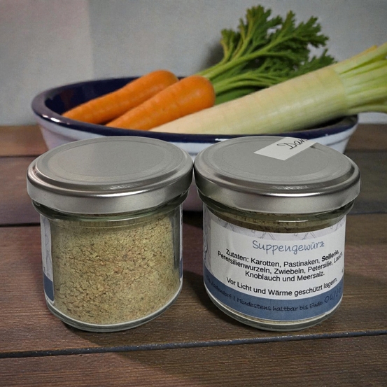 Zwei Glasgläser mit Deckeln, beschriftet mit 'Suppengewurz', stehen auf einer Holzoberfläche mit einer Schüssel mit Karotten und Petersilie dahinter.