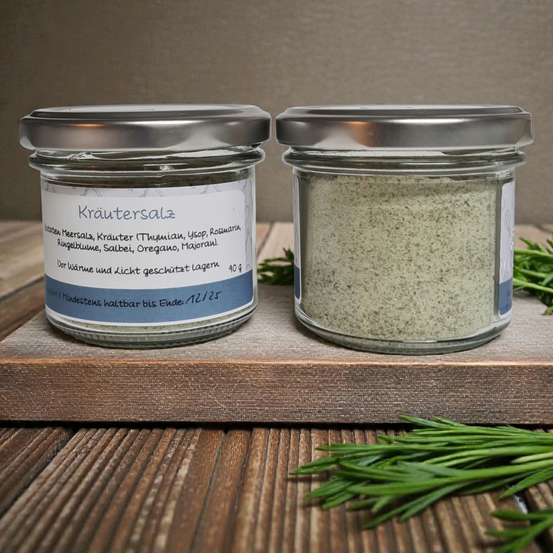 Zwei Glasgläser mit silbernen Deckeln auf einem Holzbrett, die weißes Pulver enthalten. Die Etiketten zeigen an, dass es sich um Kräutersalze handelt, mit in deutscher Sprache aufgeführten Zutaten.
