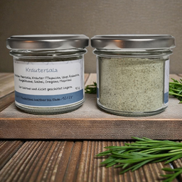 Zwei Glasgläser mit silbernen Deckeln auf einem Holzbrett, die weißes Pulver enthalten. Die Etiketten zeigen an, dass es sich um Kräutersalze handelt, mit in deutscher Sprache aufgeführten Zutaten.