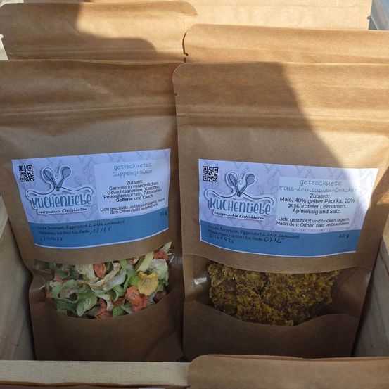 Zwei braune Papiertüten gefüllt mit verschiedenen trockenen Zutaten. Jeder Beutel hat ein blaues Etikett mit Text und einem QR-Code. Die linke Tüte enthält grünes Gemüse und die rechte Tüte enthält gelbe Kräuter.