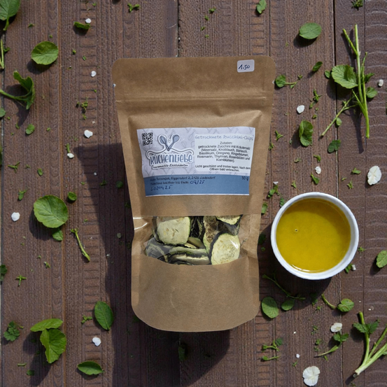 Ein brauner Beutel mit 'Kuchenliebe' Zucchini-Chips auf einem Holztisch. Daneben steht ein weißer Becher mit einer gelblichen Flüssigkeit, und um ihn herum sind grüne Blätter verstreut.
