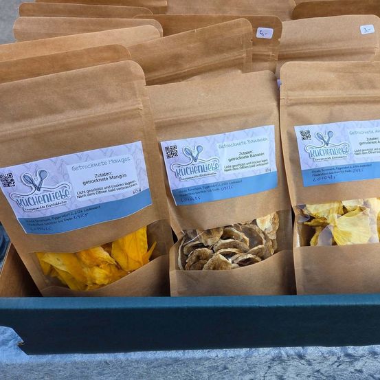 Ein Tablett mit mehreren braunen Tüten mit getrockneten Früchten, einschließlich Mangos und Bananen. Jede Tüte ist mit dem Produktnamen und einem QR-Code beschriftet.