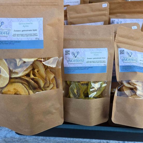 Mehrere Beutel mit getrockneten Früchten, einschließlich Äpfel und Kiwis, sind ausgestellt. Die Beutel sind braun mit blauen Etiketten. Die Etiketten zeigen die Inhaltsstoffe und das Gewicht der Produkte an.