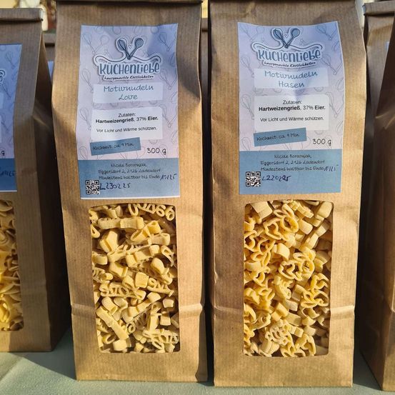 Drei braune Papiertüten enthalten Makkaroni und Käse. Jede Tüte hat ein Etikett mit 'Kuchenliebe' und 'Motivnudeln Love'. Die Tüten sind gefüllt mit Makkaroni und haben einen QR-Code. Die Makkaroni sind goldgelb.