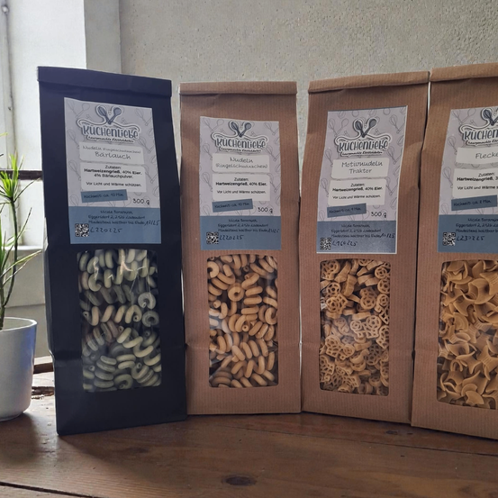 Vier Nudelpakete in verschiedenen Formen stehen auf einem Holztisch. Jedes Paket ist mit dem Markennamen und den Produktdetails beschriftet. Die Nudeln sind in Plastik verpackt.