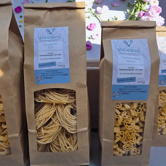 Zwei braune Papiertüten mit sichtbarer Pasta durch das Fenster. Die linke Tüte hat 'Spaghetti Nr. 5' darauf geschrieben. Die rechte Tüte hat 'Motivnudeln Herzen' darauf geschrieben. Blumen sind im Hintergrund.