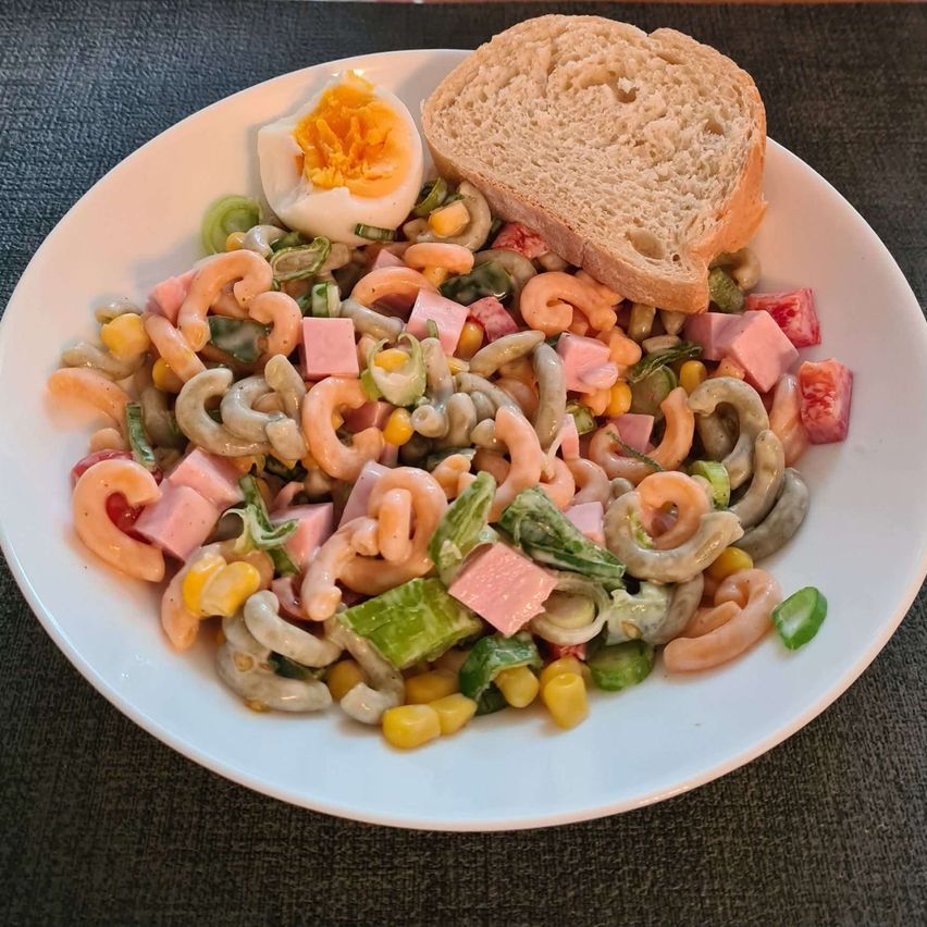 Ein weißer Teller gefüllt mit Nudelsalat, überzogen mit einer Scheibe Brot und einem halbgekochten Ei. Der Salat besteht aus Nudeln, gewürfelten Tomaten, Mais, grünen Bohnen und Schinkenstücken.